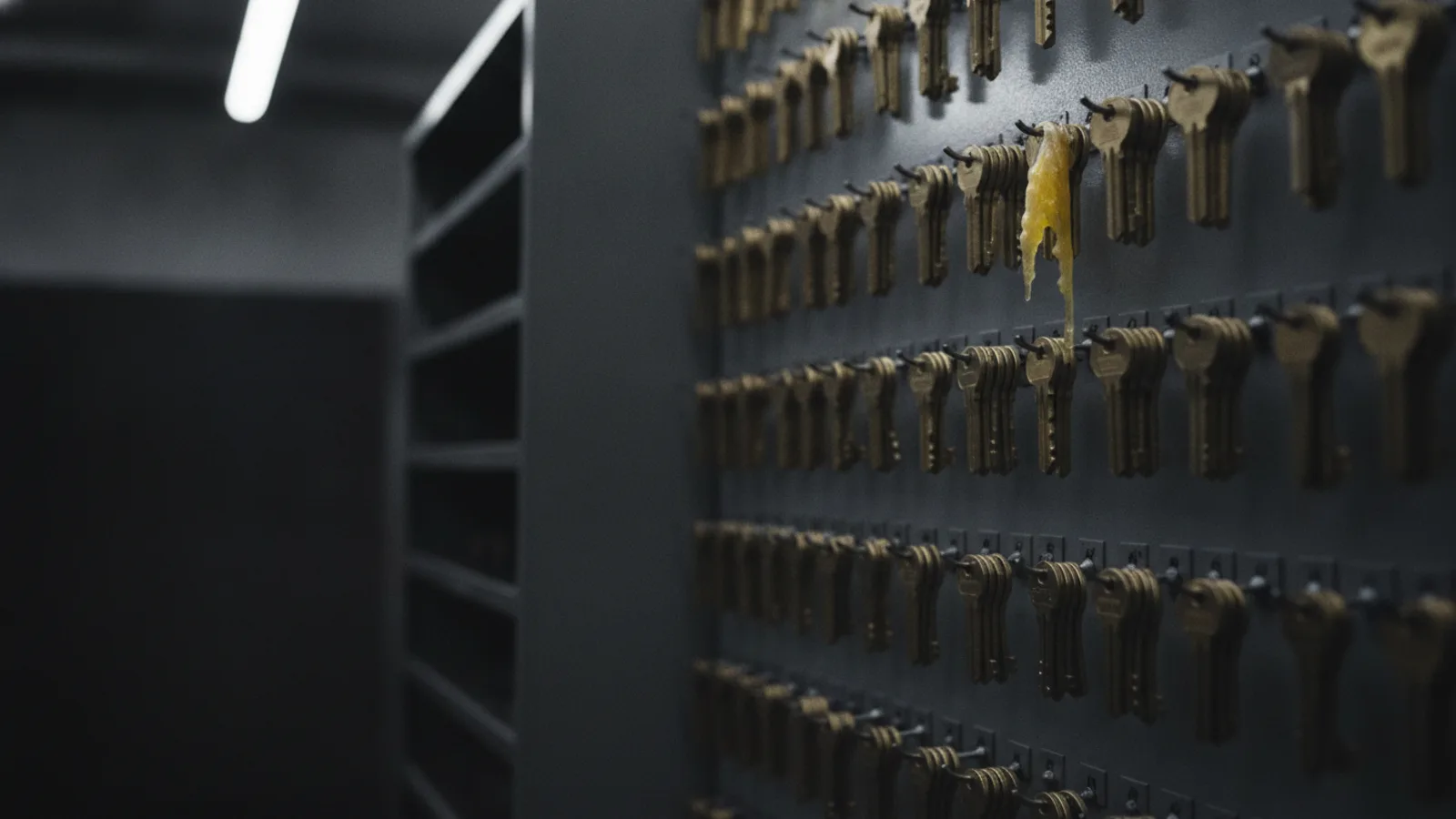 A board of identical brass keys hanging from numbered hooks, one key made of translucent wax drooping under its own weight.