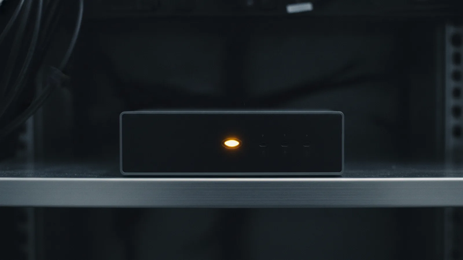 A small dark NAS enclosure on a metal shelf, one amber status LED glowing alone on its faceplate, the rest of the front panel in darkness.