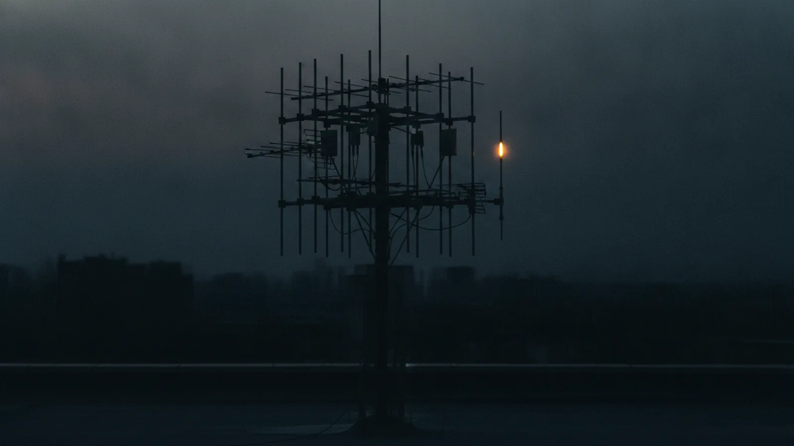 Une grappe d’antennes radio métalliques sombres plantées sur un mât de toit au crépuscule, une seule extrémité d’antenne éclairée d’une lueur ambre stable.